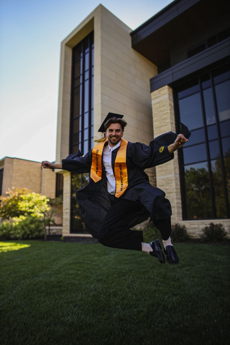 A graduate in cap and gown mid-jump on the lawn in front of a modern campus building