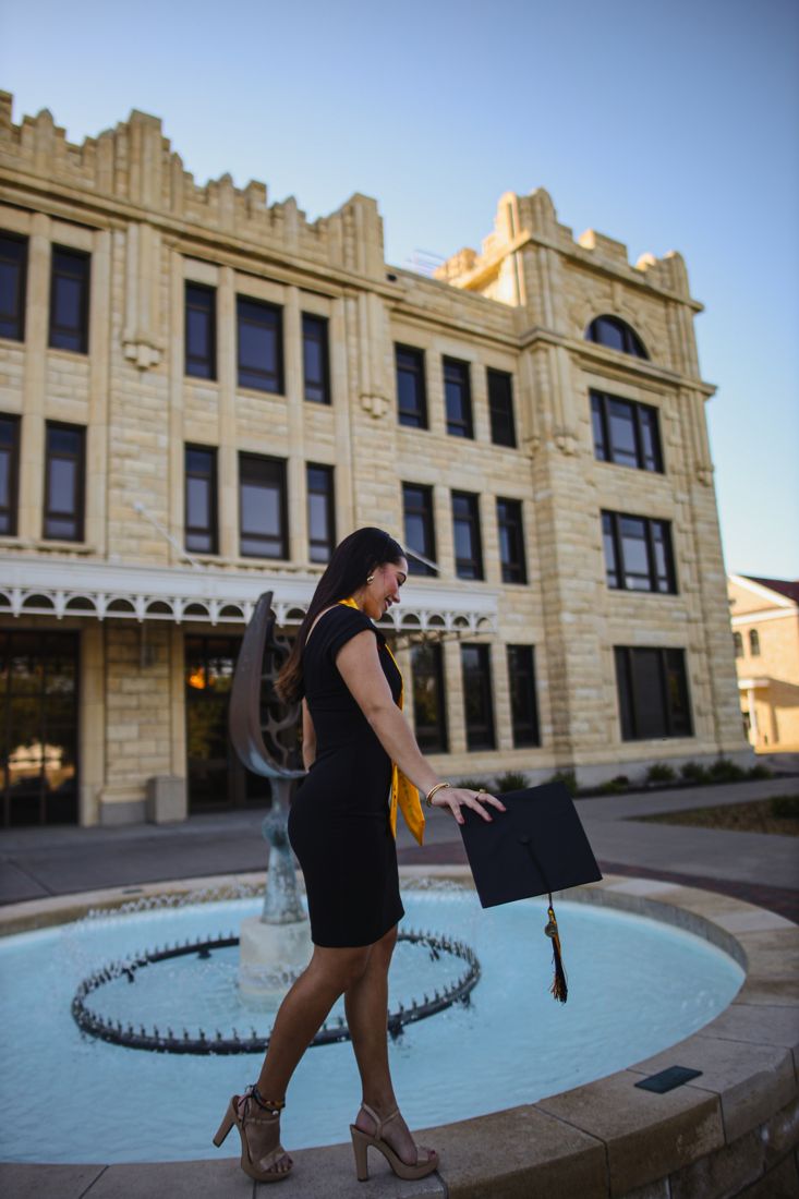 A graduate in a black dress holding her cap beside a fountain in front of a historic campus building