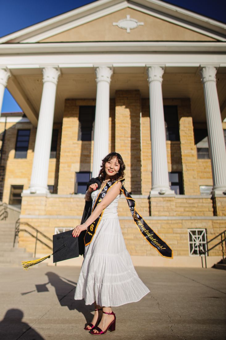 A graduate in a white dress twirling with her cap on the steps of a columned building