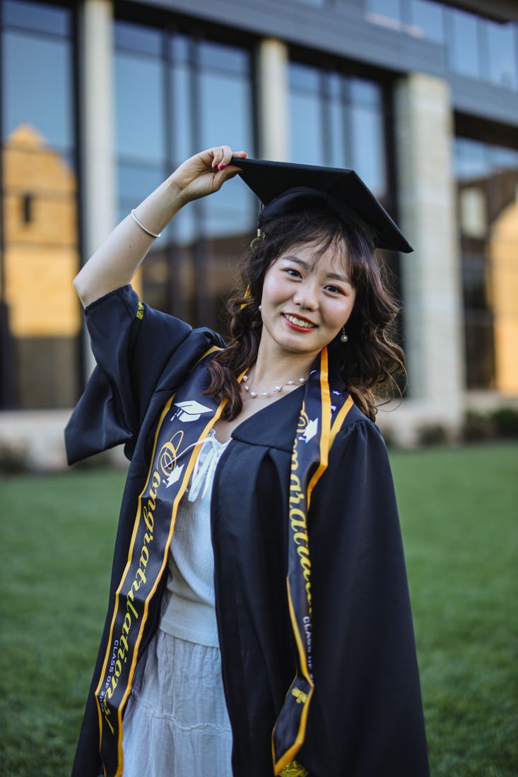 A smiling graduate in cap and gown with one hand on her cap