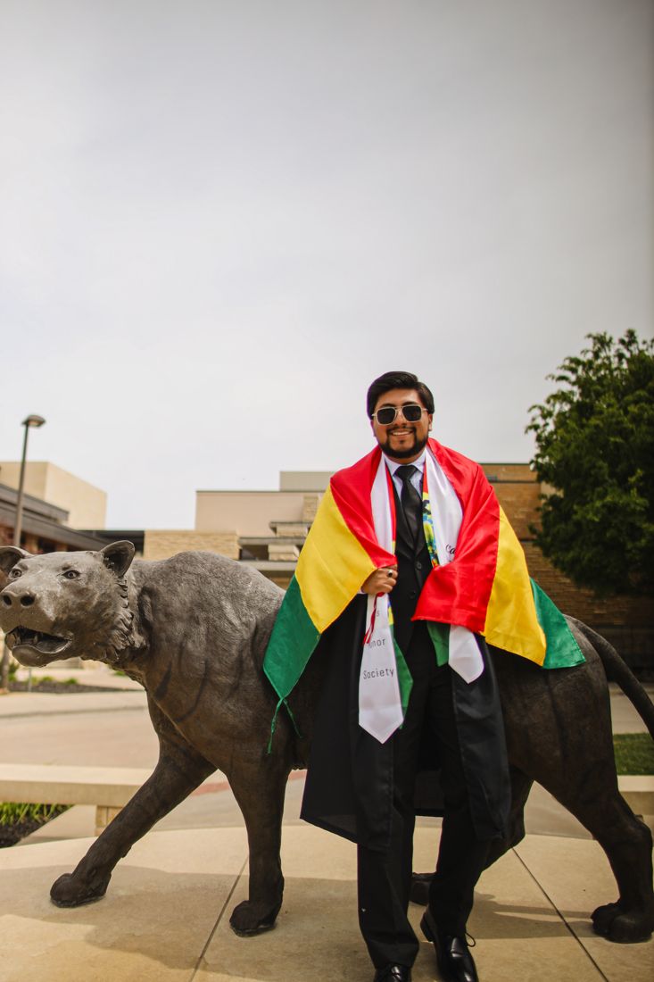 An international graduate wrapped in his country's flag beside the bronze tiger statue on campus