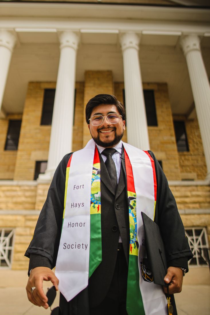 A graduate in an Honor Society stole holding his cap in front of a columned building